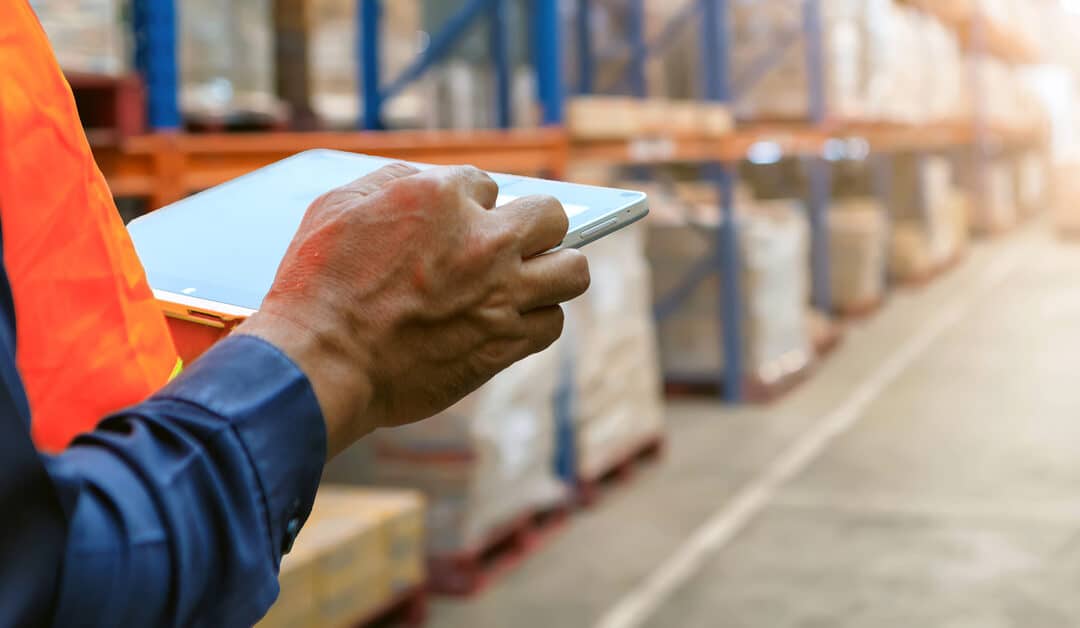 An employee in an orange safety vest stands in a fully stocked warehouse checking its inventory on a tablet.