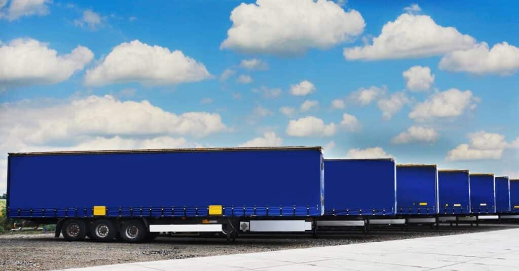 A lineup of seven blue storage trailers are next to each other underneath a blue sky with many clouds.