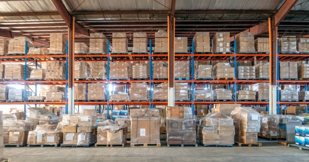 The interior of a neatly organized warehouse shows cardboard boxes wrapped in plastic wrap and sitting on palettes.