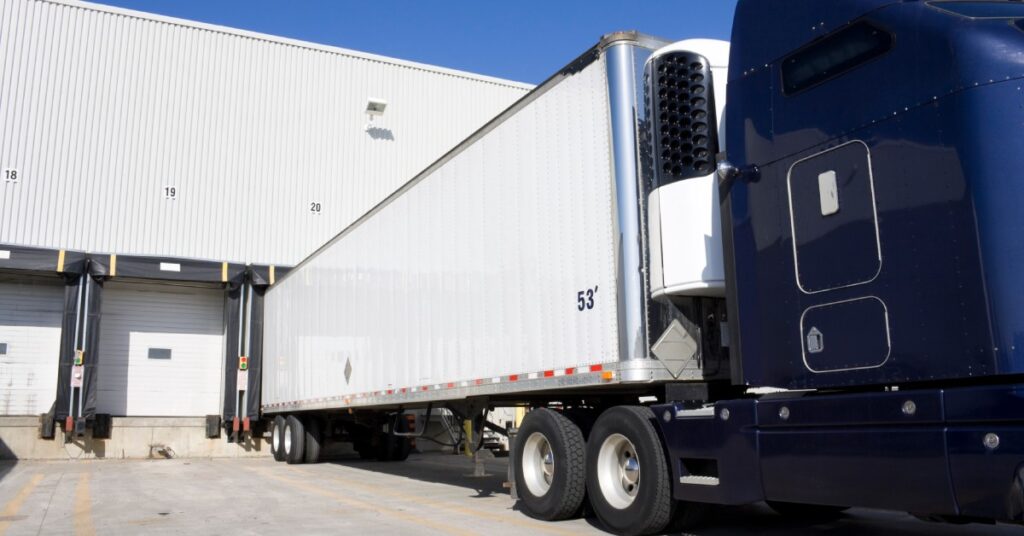 A blue transport truck backs up its storage trailer into a warehouse. There are several other open garages nearby.