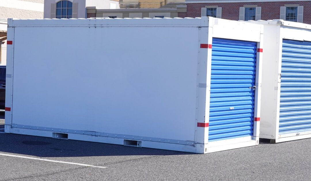 Two large white storage containers with blue doors. They sit side by side in a business parking lot.