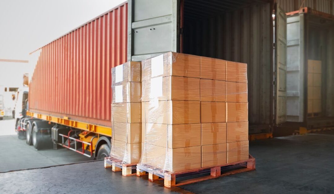 A pallet of cardboard boxes sits, waiting for a worker to load it into a storage trailer in a business warehouse.