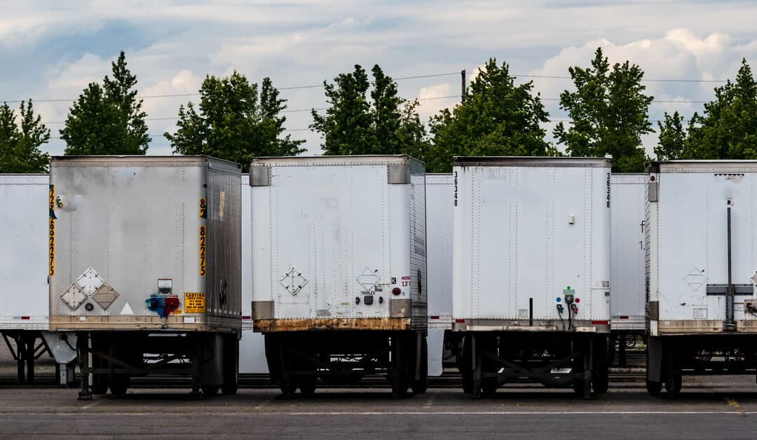 A row of storage trailers on vehicles lined up in a parking lot. Four white trailers with their access doors shut.