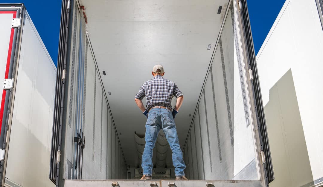 A man stands inside an empty semi-truck trailer. He wears a ball cap, a plaid shirt, and blue jeans.