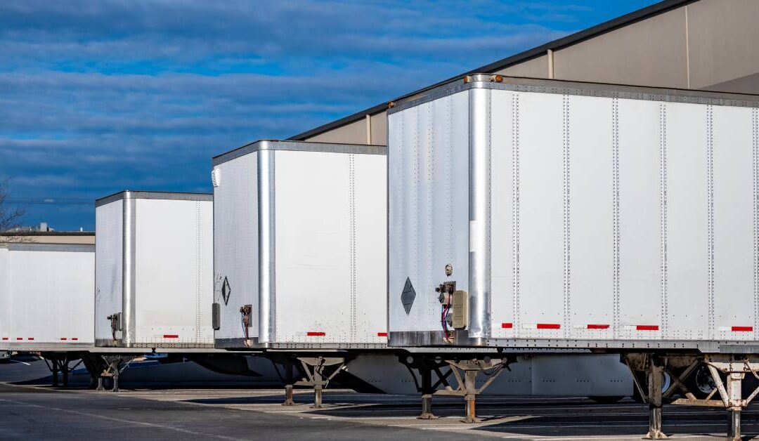 Several white semi-trailers stand in a line alongside a building. The sky in the background is blue.