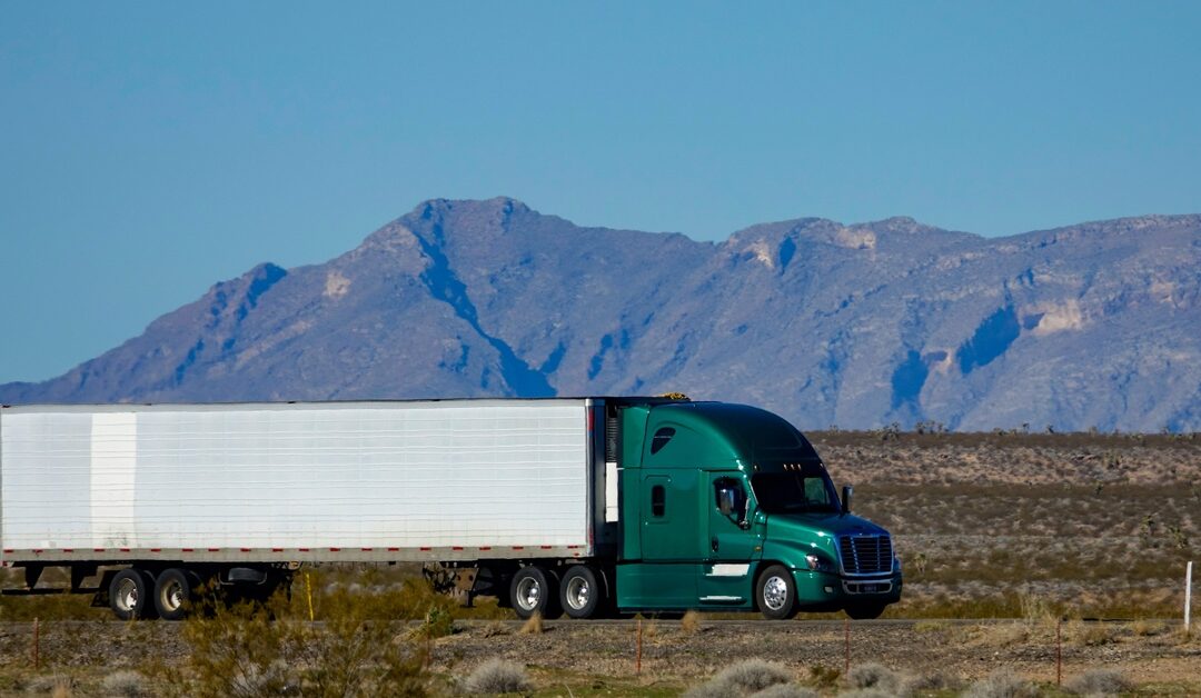 A green semi-truck tows a white semi-trailer as it drives through a barren area. Mountains are in the background.