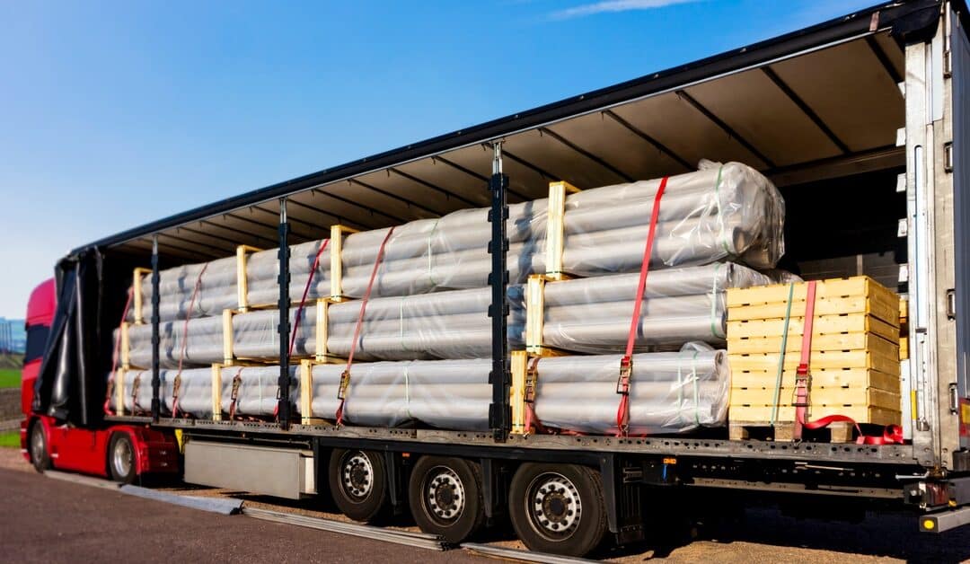 A red truck with an open semi-trailer is loaded with metal tubes and wooden pallets while parked under a blue sky.