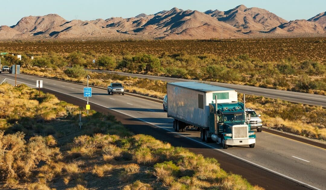 A sleek, green American truck drives on the interstate highway, behind him are traffic, surrounded by grasslands.