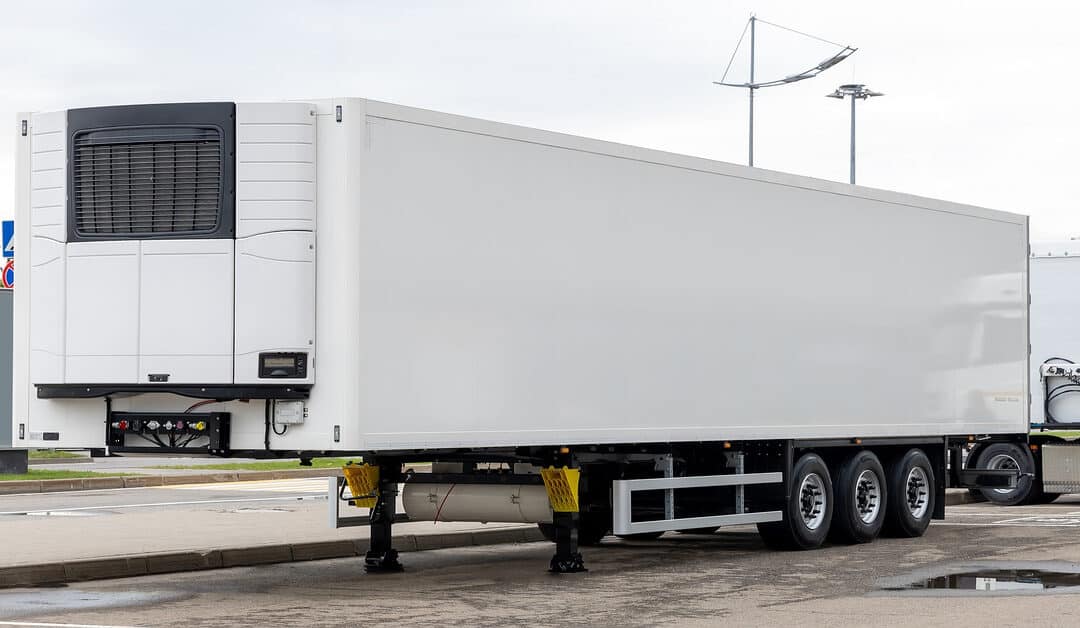 A sleek, white, refrigerated semi-trailer is parked on concrete, surrounded by puddles of water.