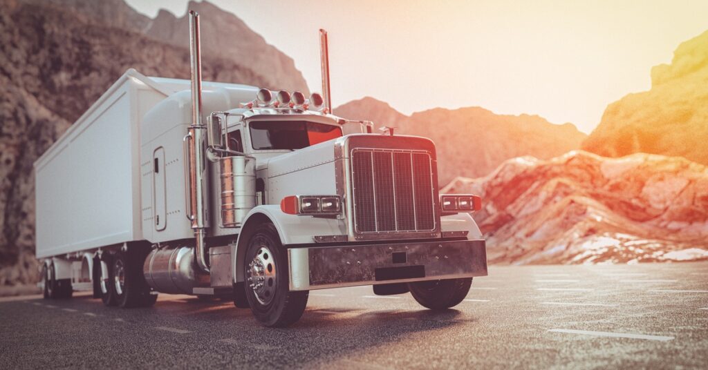 A white American-style semi-trailer truck with a load drives on the road. Behind it are mountain ridges.
