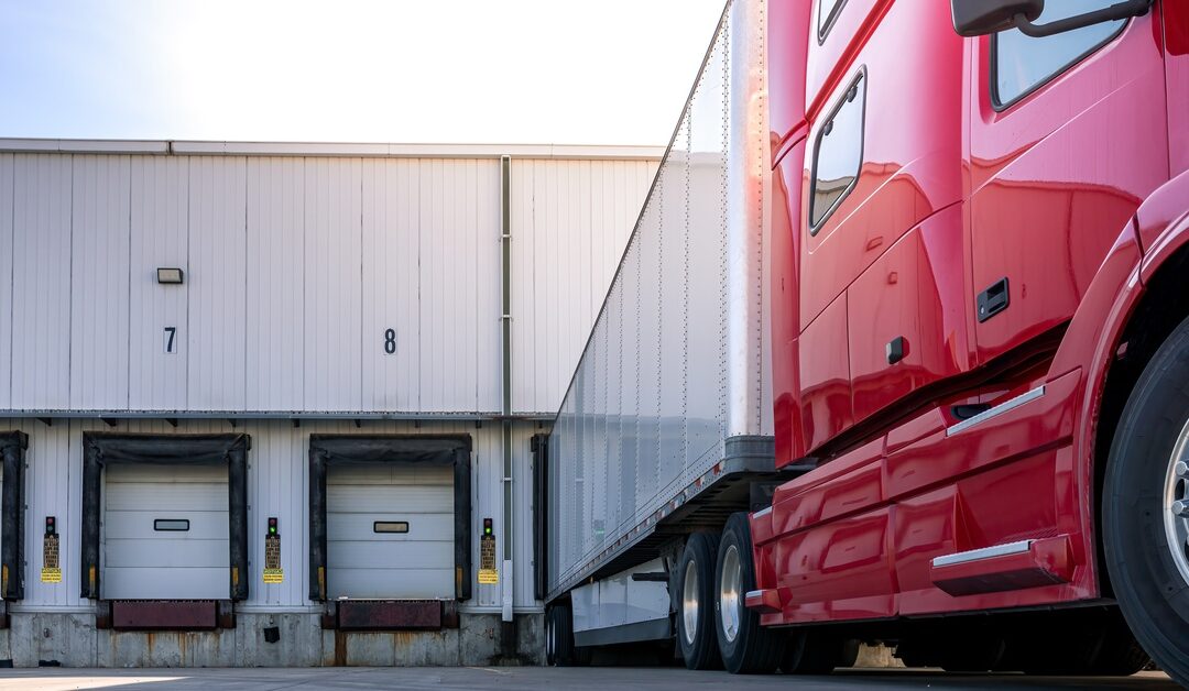 A modern, red American truck with a cargo trailer parks at the docks, waiting for a load, underneath a clear sky.