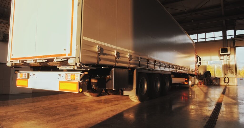 A semi-trailer truck with a cargo load pulls into a service station with sleek, epoxy flooring for maintenance.
