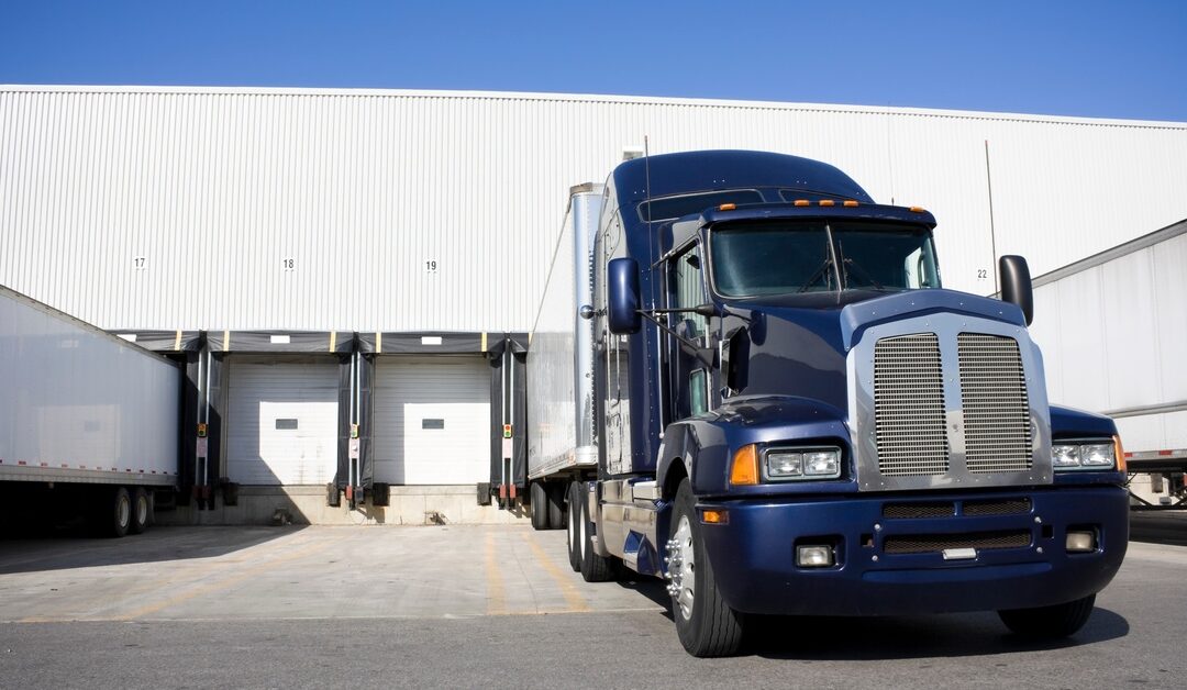 The sun shines down on a dark blue semi-trailer truck as it docks right in front of a white warehouse building.