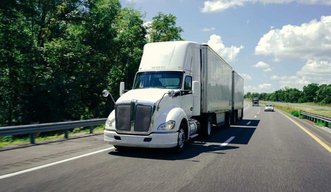 A white semi-truck with double trailers attached to it drives down the highway, with two vehicles behind it.