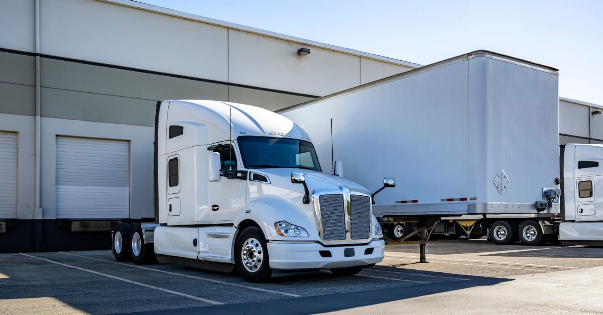 A white semi truck parks next to a dry van trailer in front of the warehouse dock gates on a sunny day.