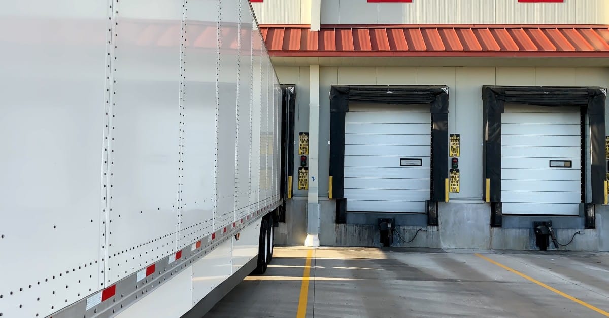 A long, 53-foot semi trailer parks next to the red and white warehouse docks to pick up or deliver a load.
