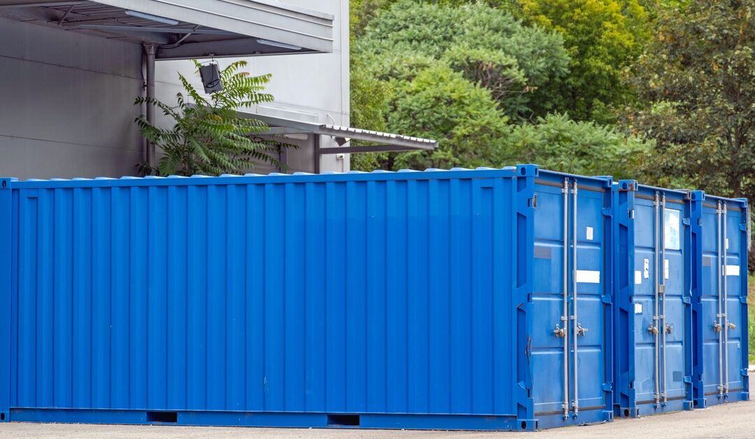 Three blue storage cargo containers sits side by side, in a row, next to a white building surrounded by trees.