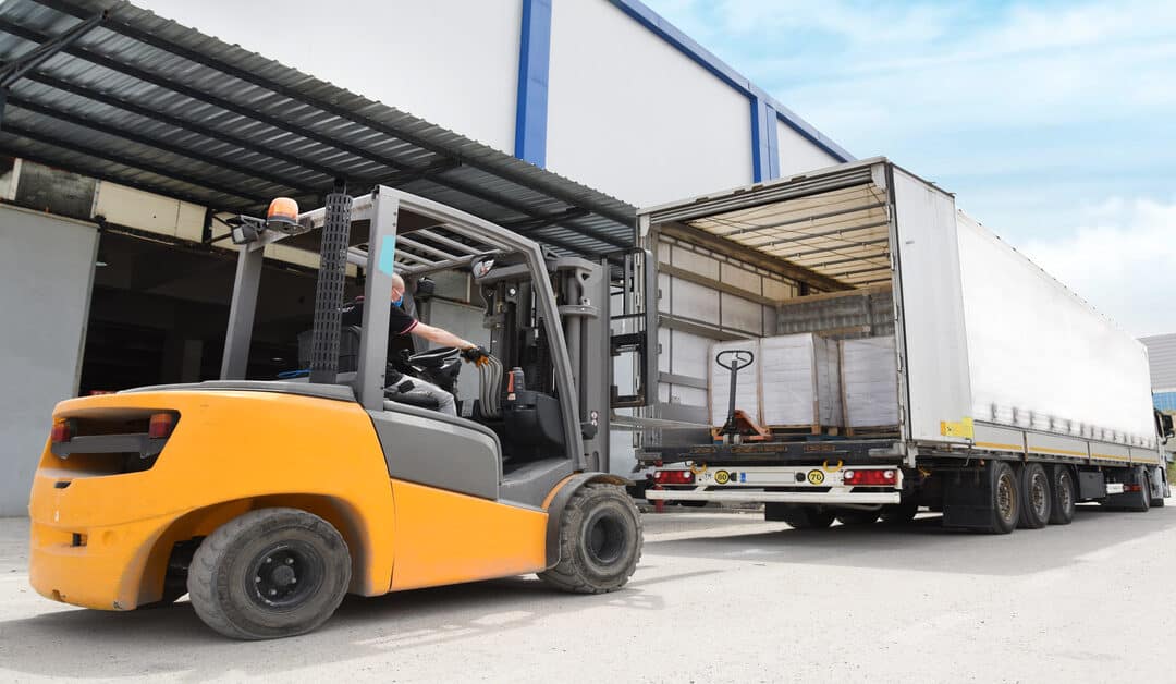 A yellow forklift loads cargo onto a white semi-trailer truck parked beside a white and blue building.
