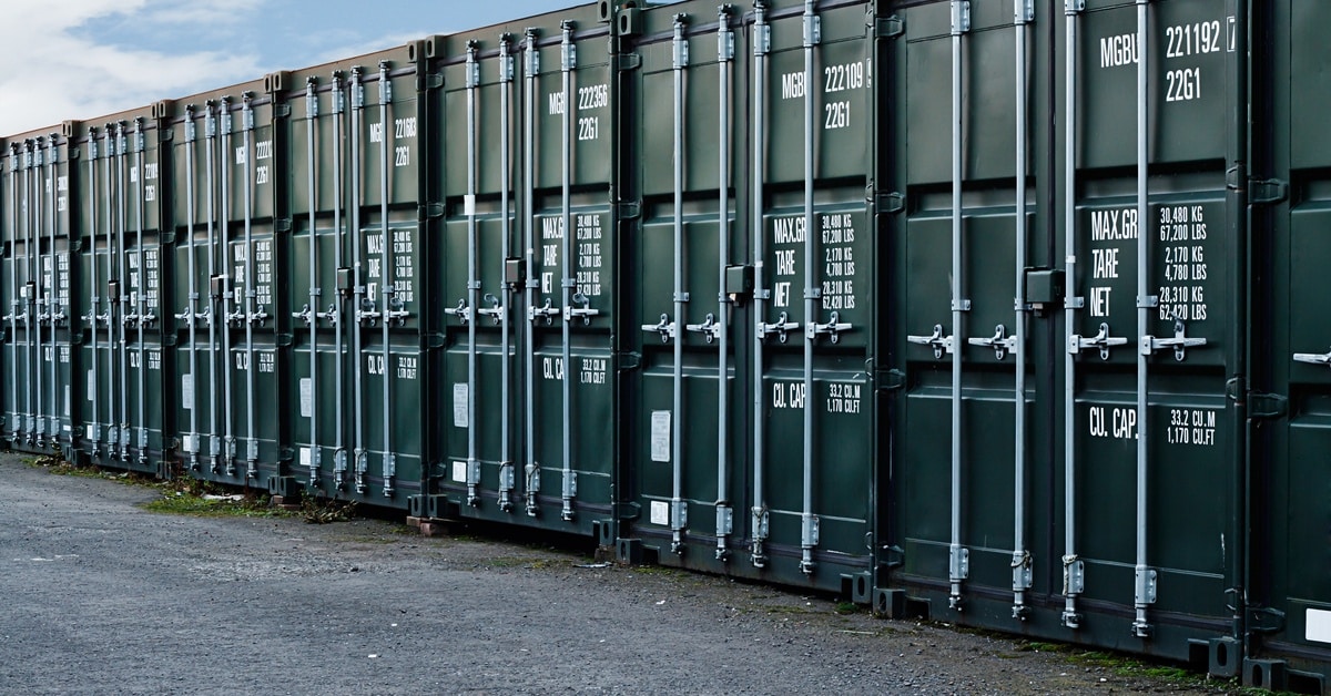 A row of dark green shipping containers with various weight labels sit on the ground under a blue sky.