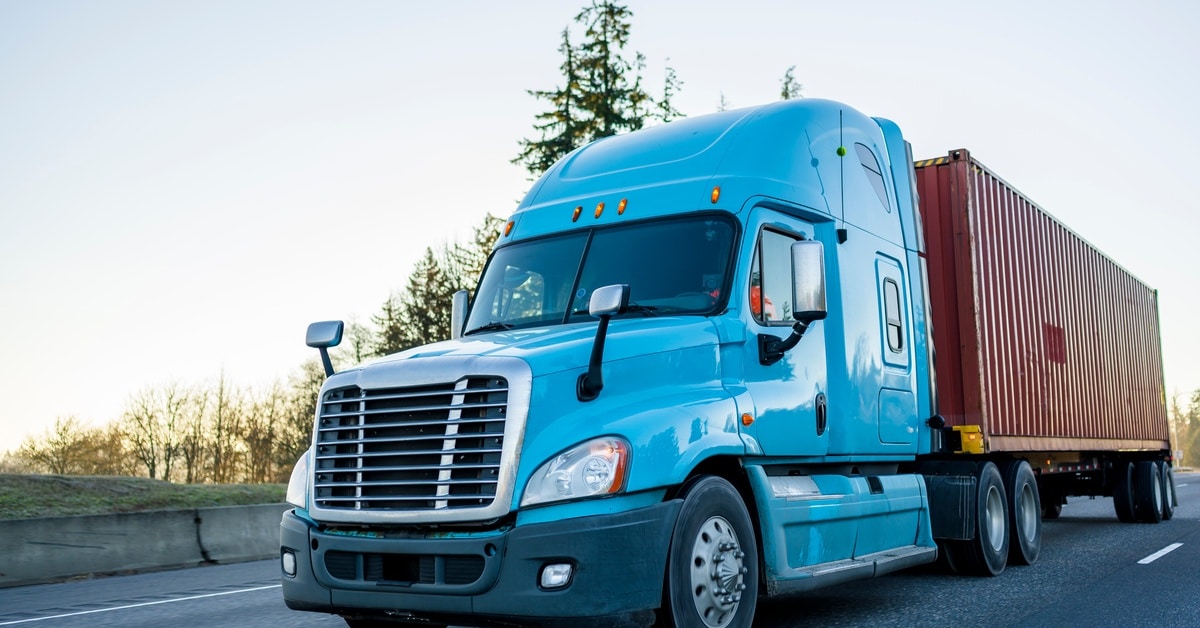A light blue semi-trailer truck with a red container load drives down the highway, next to a row of trees.