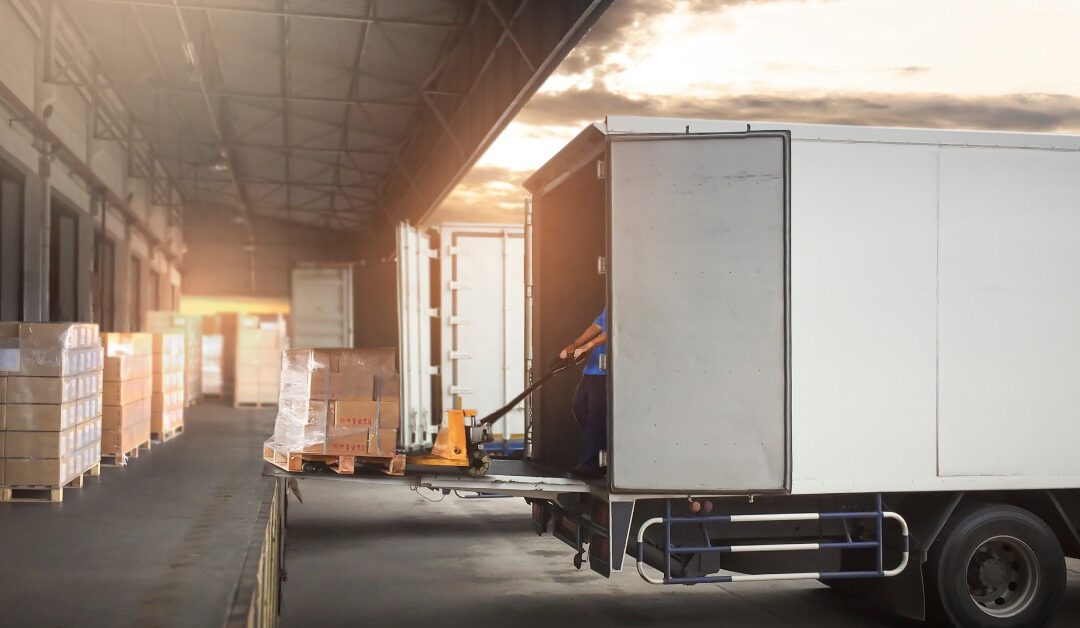 A worker hauls a pallet and loads it into a cargo trailer parked in front of a warehouse loading dock.