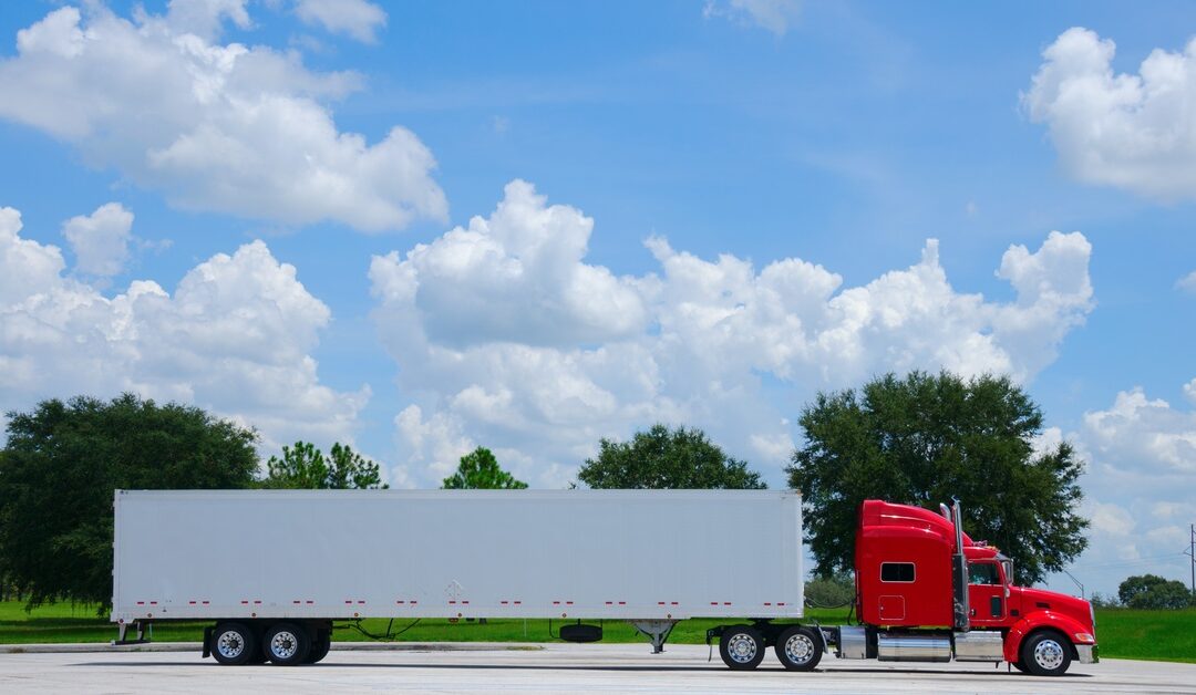 A red 18-wheel semi-truck with a white cargo trailer parks on the road in front of trees, under a clear blue sky.