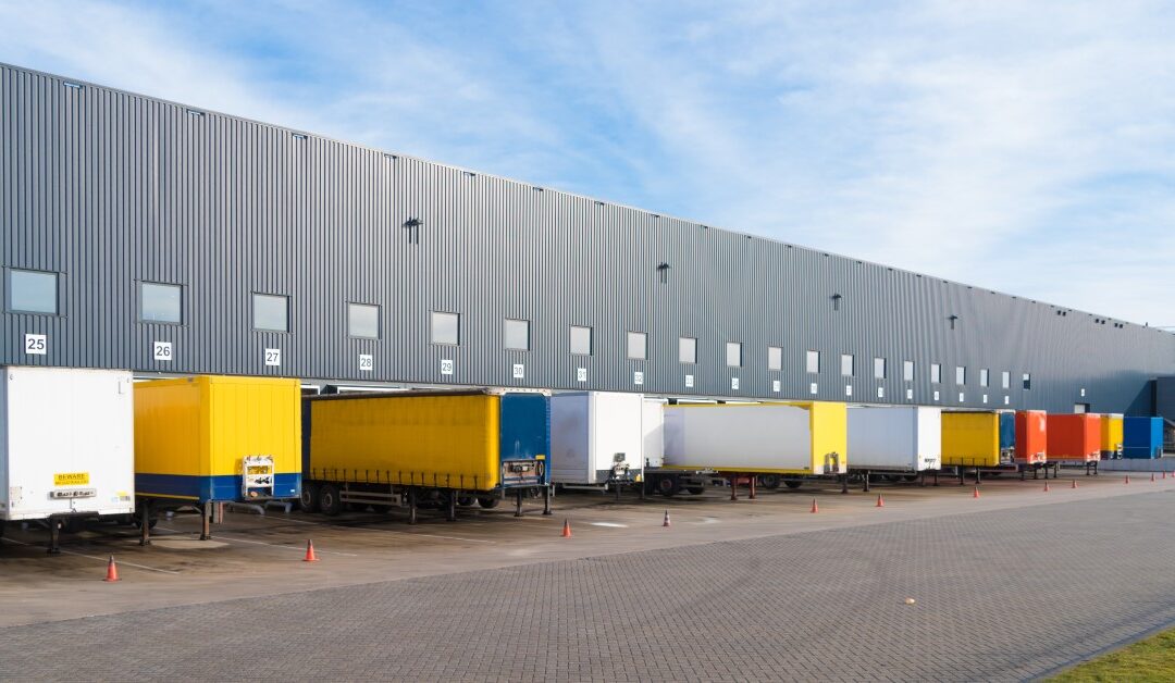 Multiple, colorful trailers line up in a row, next to traffic cones, in front of a commercial warehouse.