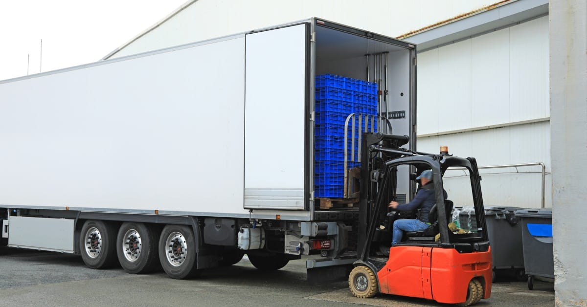 A worker uses an orange forklift to load a stack of blue crates into a parked white semi-trailer truck.