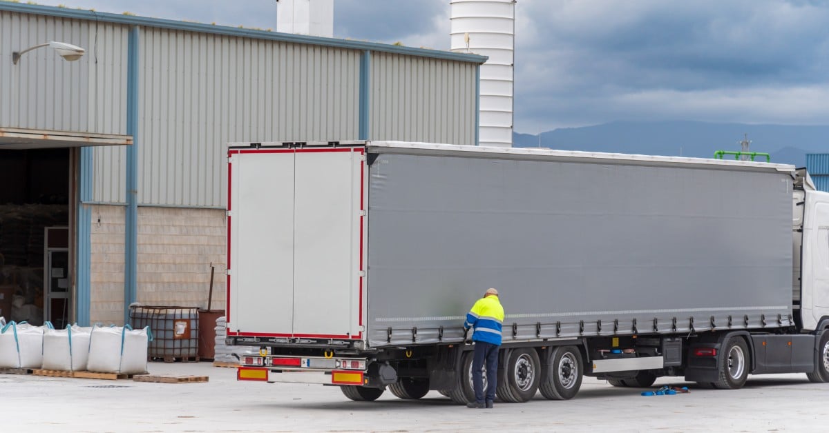 A worker wearing a high-vis vest closes the side of a white semi-trailer parked beside a warehouse under a cloudy sky.