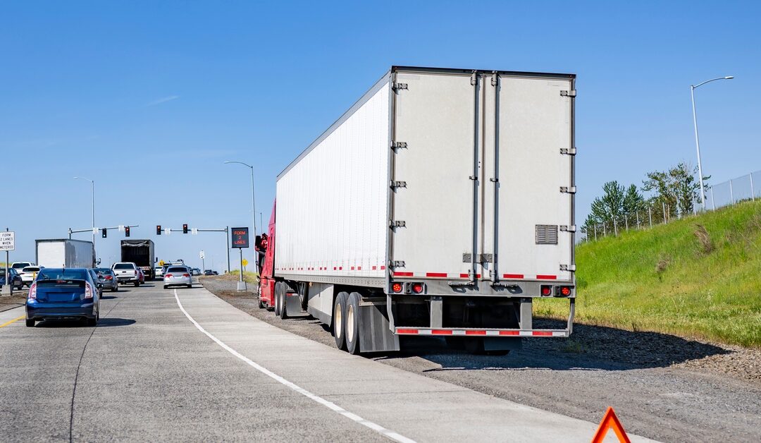 A red semi-trailer truck breaks down on the highway, with an emergency triangle behind it, underneath a clear sky.
