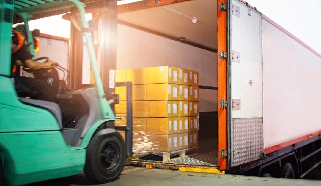 A teal colored forklift tractor loads up multiple package boxes into a container trailer parked by a warehouse.
