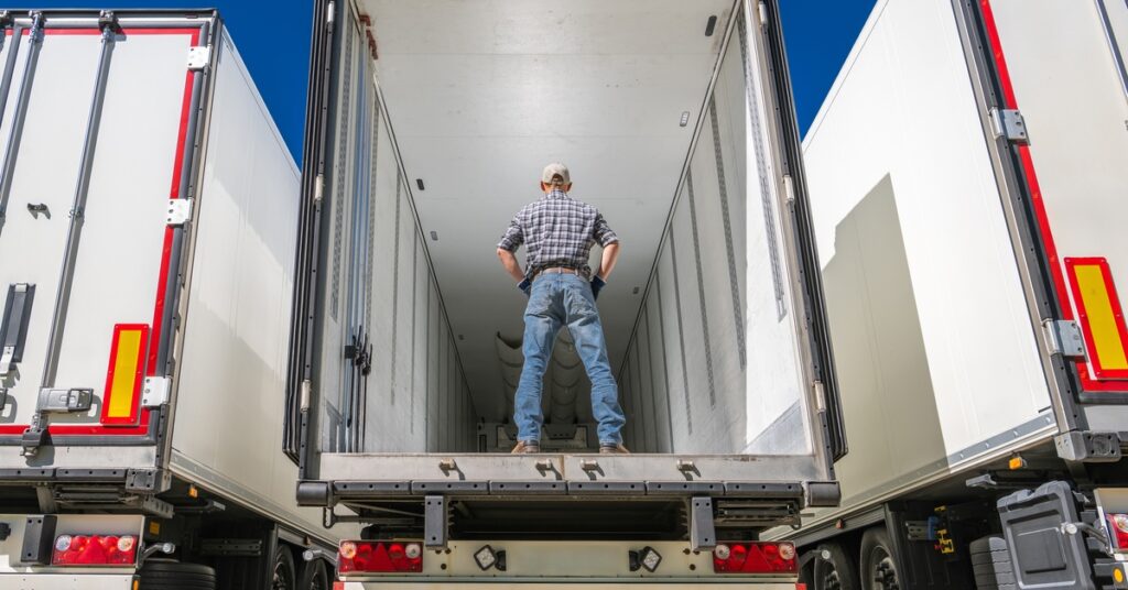 A man wearing a plaid shirt and jeans stands inside an empty semi-trailer, inspecting its interior.
