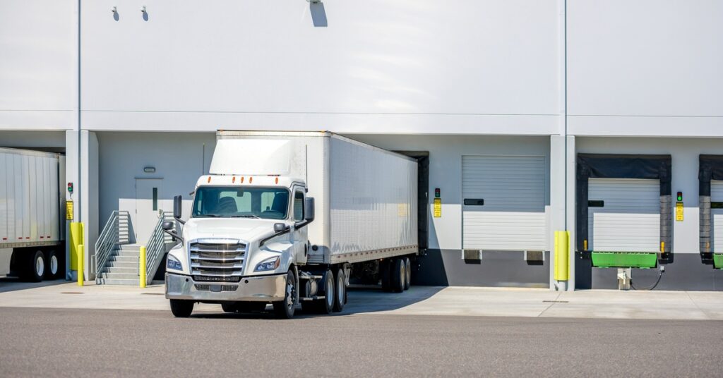 The sun reflects off a white semi-trailer truck, casting a shadow as it loads cargo at the docking gate.