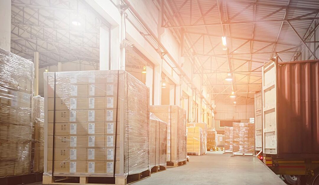 Pallets of boxed goods stacked on a loading dock beside a parked cargo container truck at a warehouse.