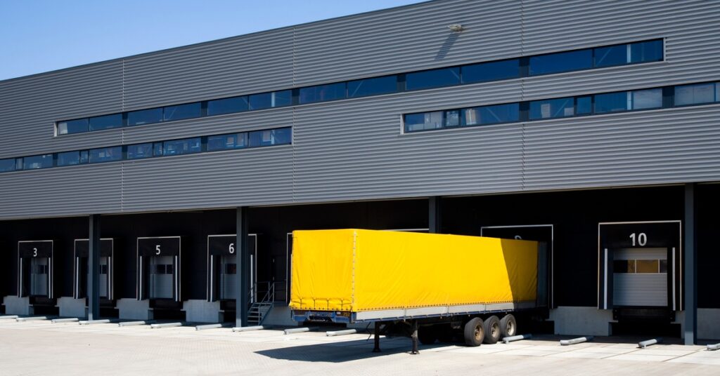 Row of numbered warehouse loading dock doors with a yellow semi-trailer backed into one bay under a clear sky.