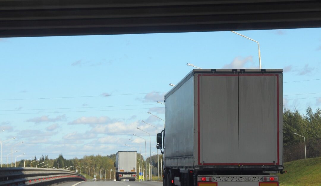 White semi-truck pulling a trailer drives under a bridge on a highway beneath a clear blue summer sky.