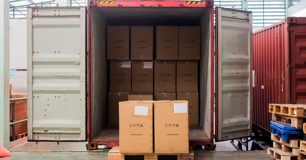 Cardboard cartons being unloaded from a shipping container, stacked near the container opening on the floor.