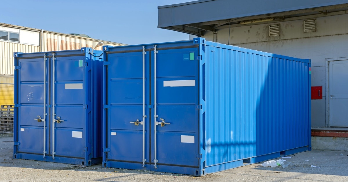 Two shipping containers placed side by side on a paved surface, with visible doors and corrugated metal walls.