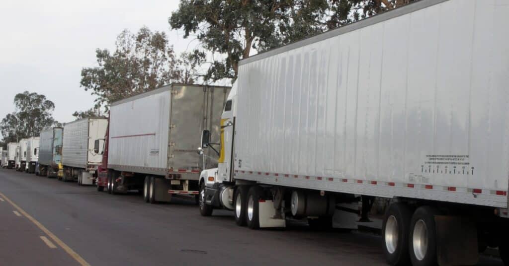 Several tractor trailers lined up along a road, with truck cabs and long trailers waiting in a row together.
