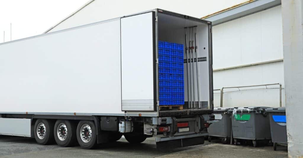A white refrigerated box truck parked beside stacked crates, with an open cargo area and people nearby.
