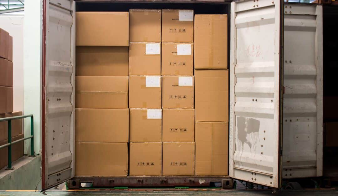 a stack of cardboard boxes into a red, open shipping container beside stacks of boxes on a spacious warehouse.