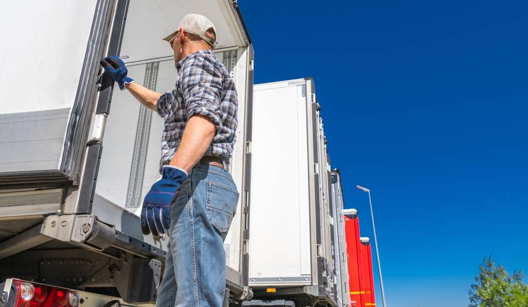 A man wearing gloves and a cap opens the rear door of a semi truck trailer parked beside other trailers.