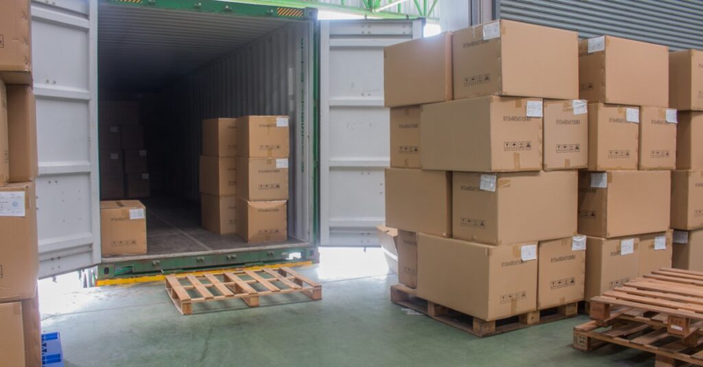 several stacks of cardboard boxes sit on wooden pallets next to an open cargo container in a warehouse.
