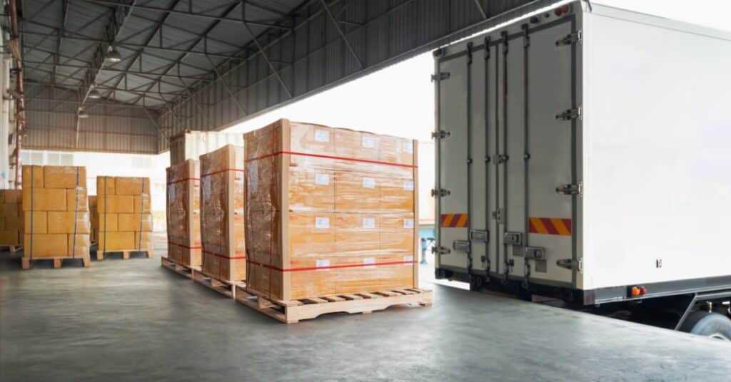 a group of wrapped cardboard boxes on wooden pallets beside a white truck in a covered loading area.
