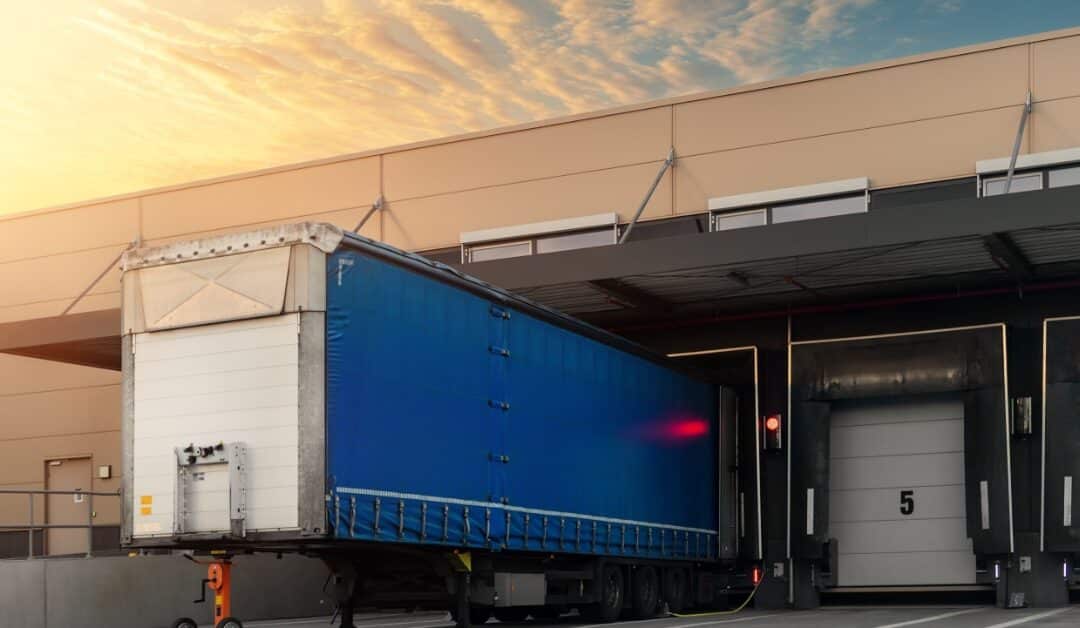 A blue semi trailer backed into a warehouse loading dock beneath a yellow and blue sky with bay doors nearby.