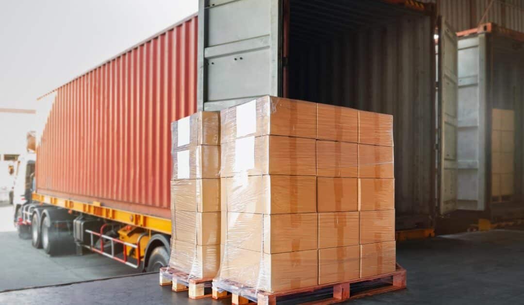 A pallet stacked with wrapped cardboard boxes beside an open shipping container at an indoor warehouse dock.