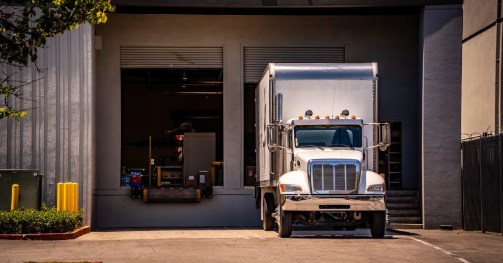 A white semi truck parked outside two loading bays at a commercial building with pavement and trees nearby.