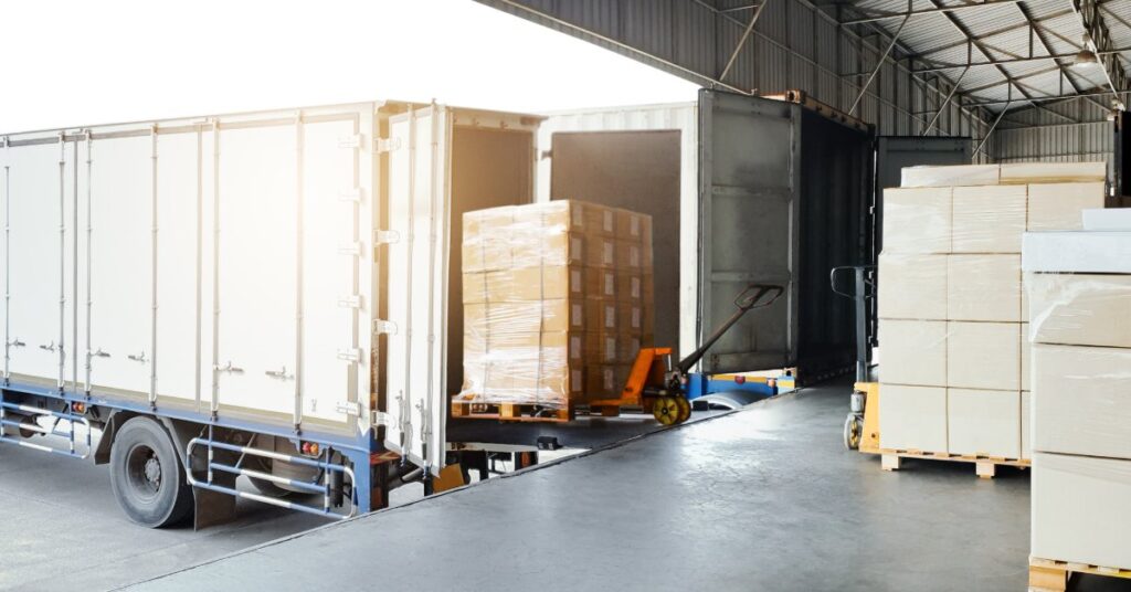 A pallet of wrapped cardboard boxes being moved from a warehouse floor into an open container truck.