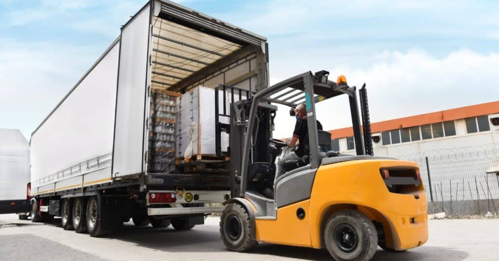 A yellow forklift loading wrapped cargo into the open back of a white trailer beside an industrial building.