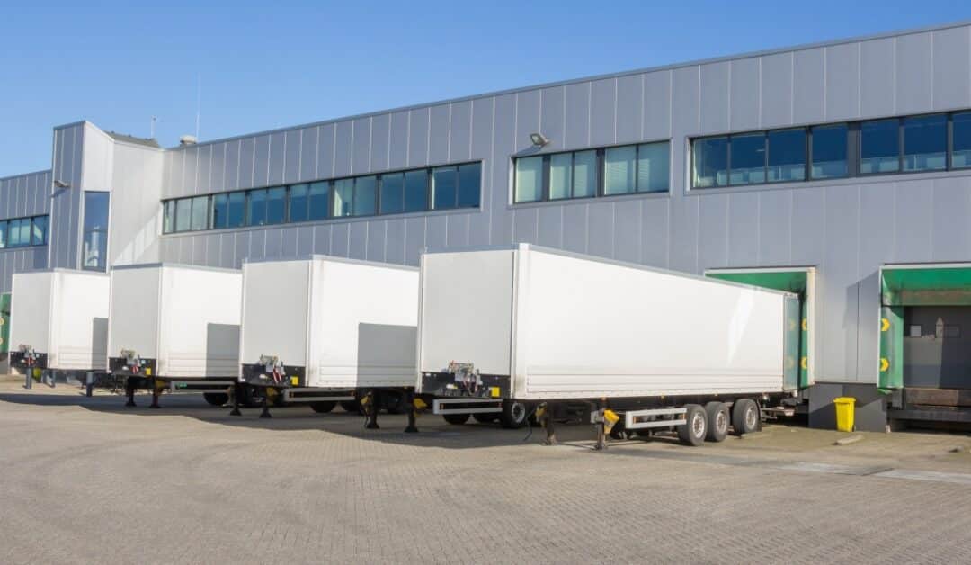A line of white trailers waiting outside a gray warehouse with green loading dock doors and paved lot.